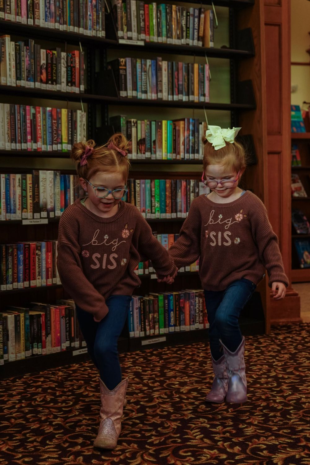 Two red-haired twin girls in matching big sis sweaters and glasses hold hands in a library.