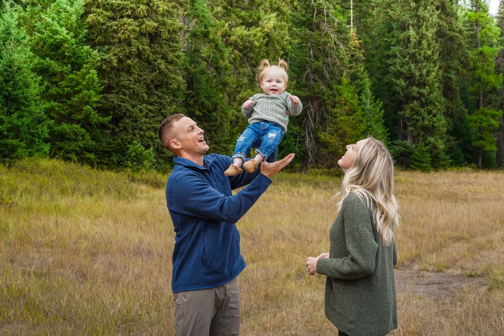 A joyful family outdoors, father lifting smiling baby in the air while mother watches, surrounded by tall green trees.