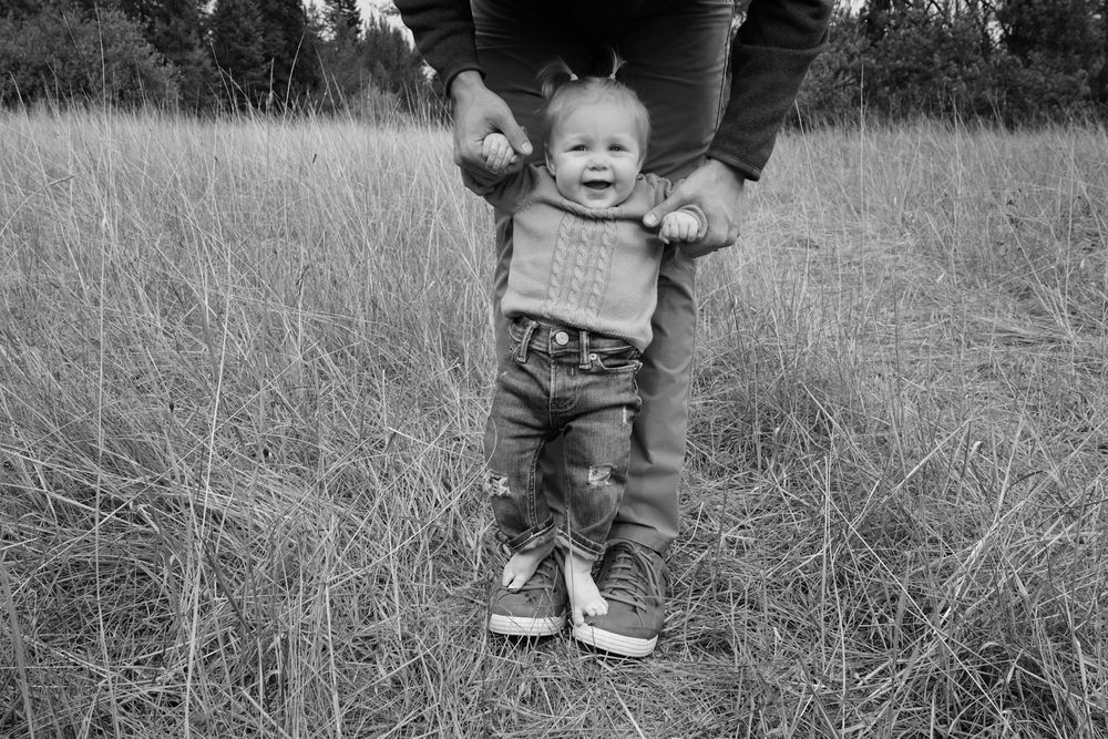 Black and white photo of a toddler learning to walk outdoors, supported by an adult in a grassy field.