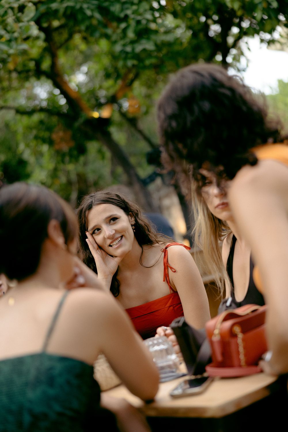A smiling woman in a red dress enjoys an outdoor dinner party with friends seated at a wooden table under trees.