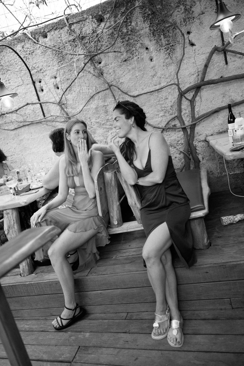 Two women in dresses laugh together on a wooden deck at an outdoor restaurant in black and white photo.