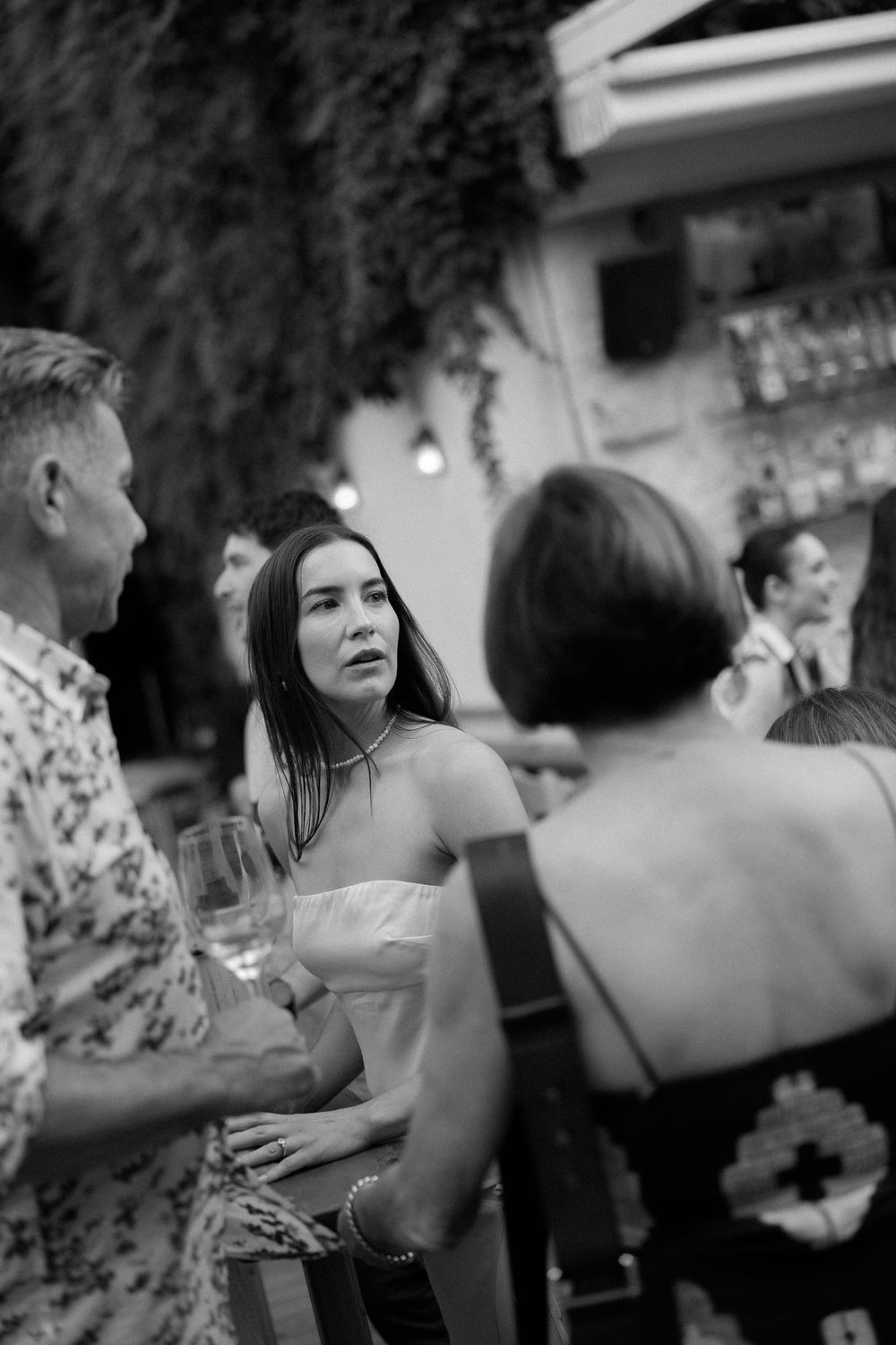 Black and white photo of guests socializing at an outdoor evening party, woman in strapless top holding wine glass.