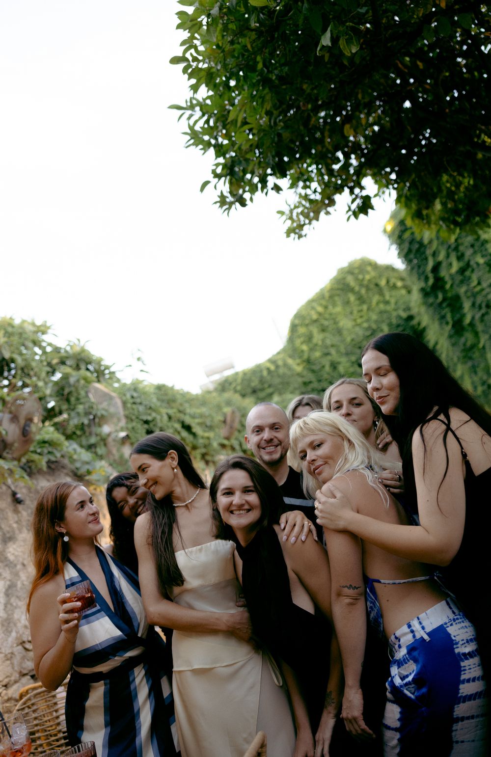A group of friends pose together outdoors near lush green ivy-covered walls at a summer gathering.