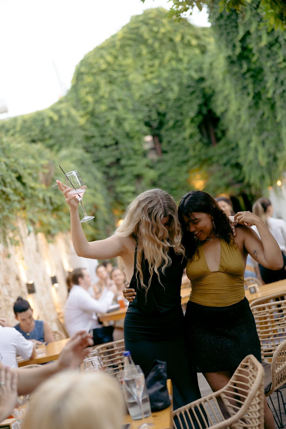 Two women dance joyfully outdoors at a garden party, one holding a champagne glass aloft.