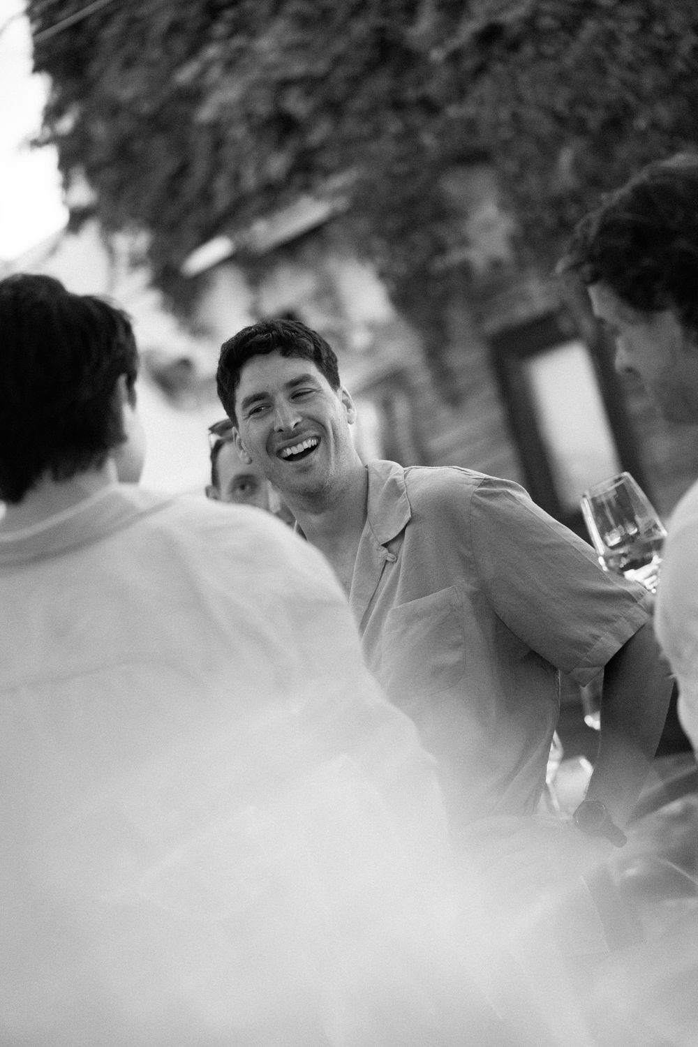 A laughing man holds a wine glass while socializing with friends at an outdoor gathering in black and white.