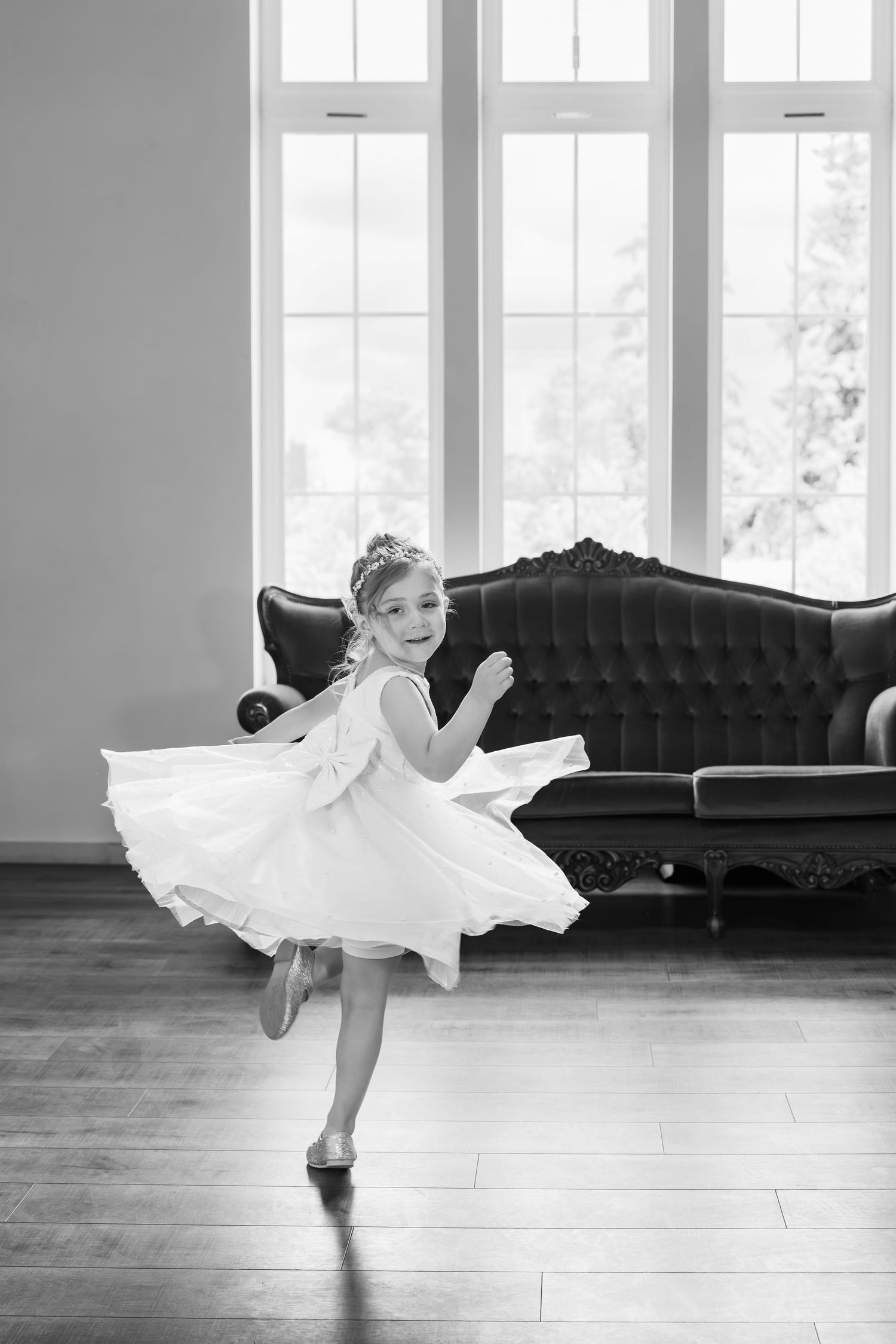 A young dancer in a white dress twirls gracefully on hardwood floors near a vintage tufted sofa by large windows.
