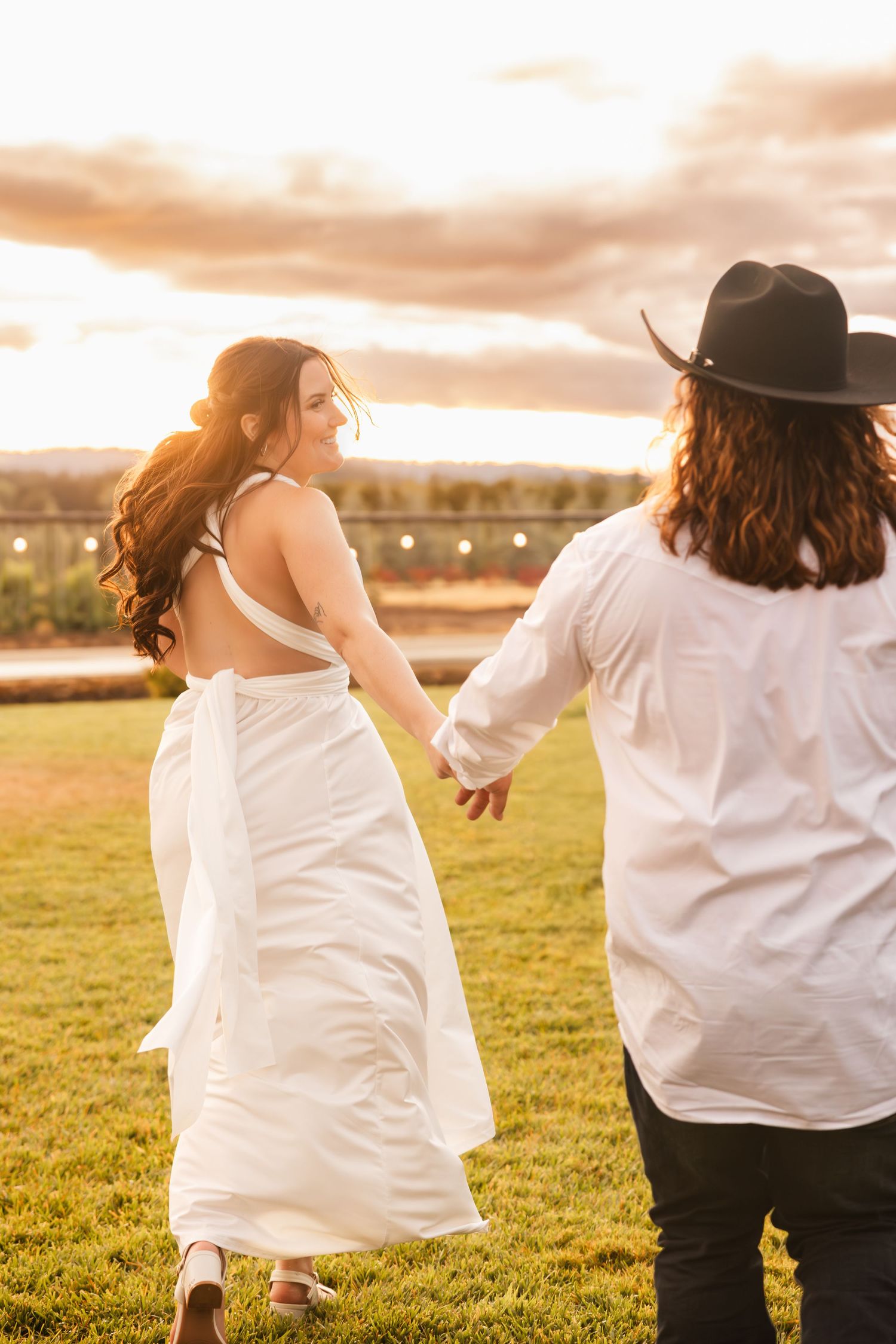 A couple holds hands and runs through a grassy field at sunset with warm golden light illuminating the scene.