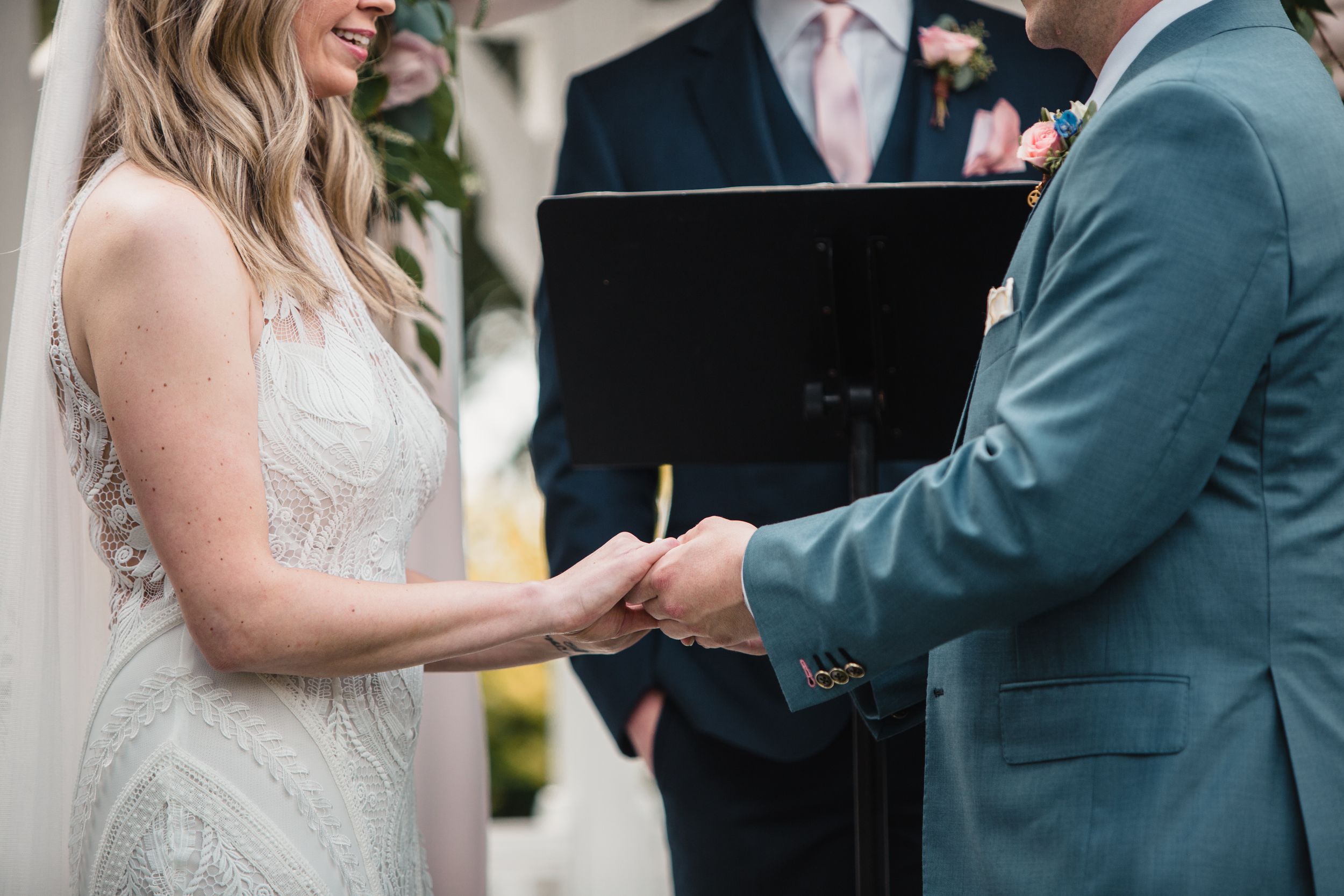 Bride and groom holding hands during outdoor wedding ceremony with officiant in background.