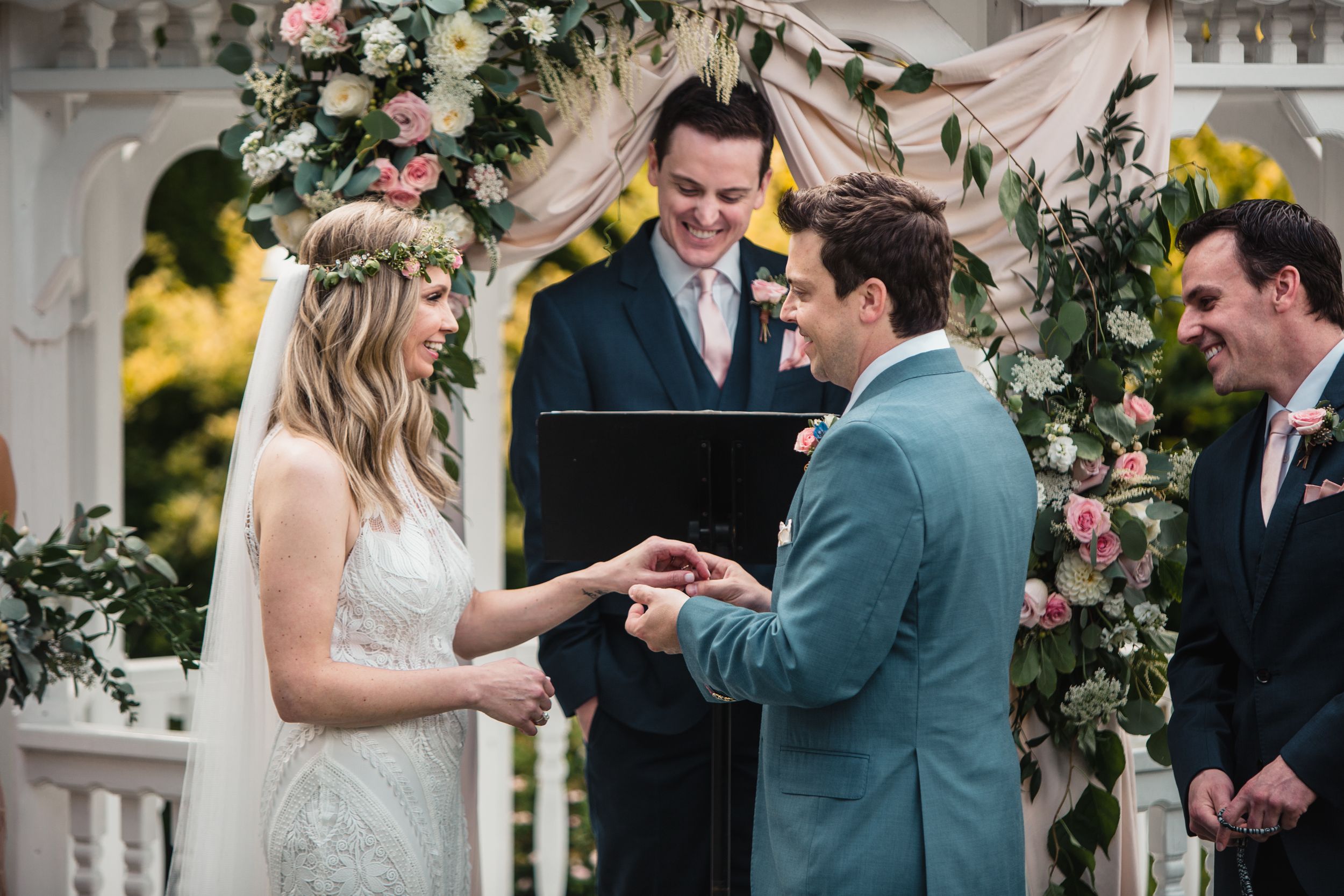 Couple exchanging rings at outdoor wedding ceremony under floral arch with officiant and groomsmen.