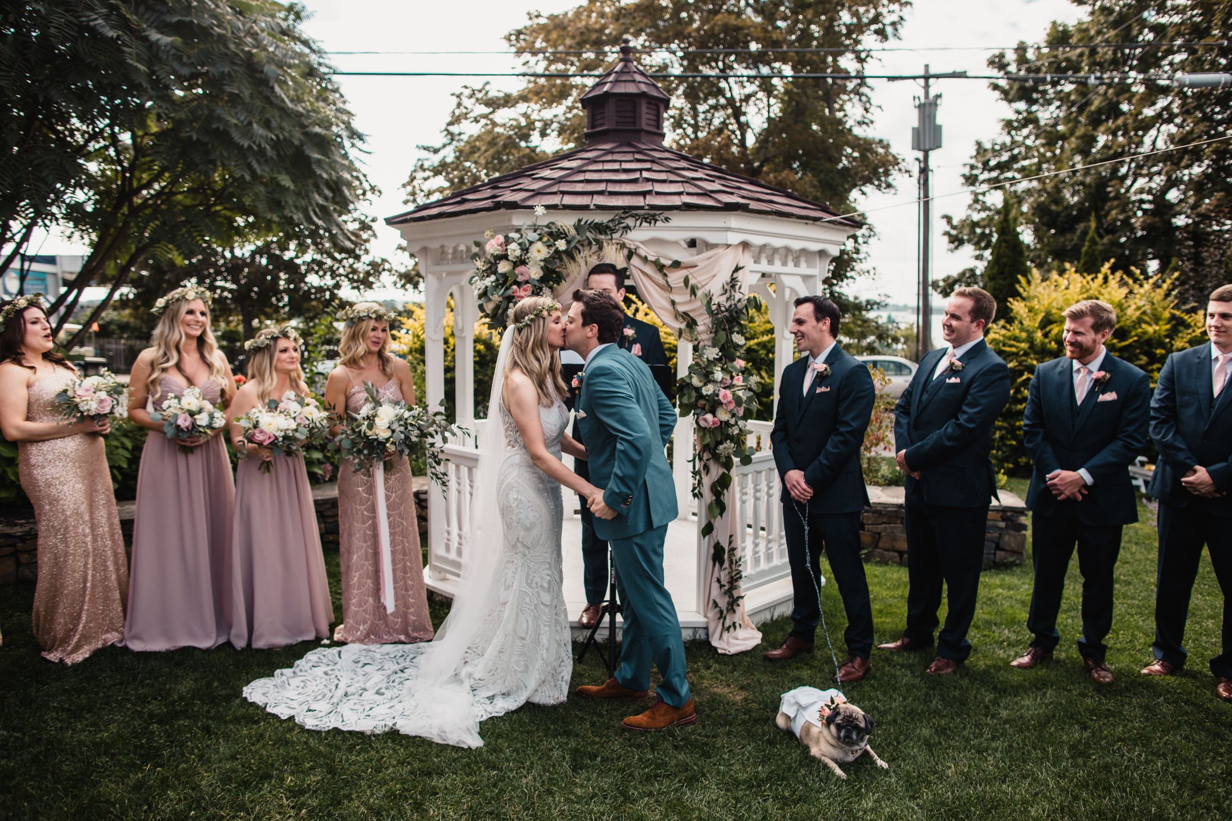 Wedding couple kissing at outdoor gazebo ceremony with bridesmaids in pink and groomsmen in navy suits.