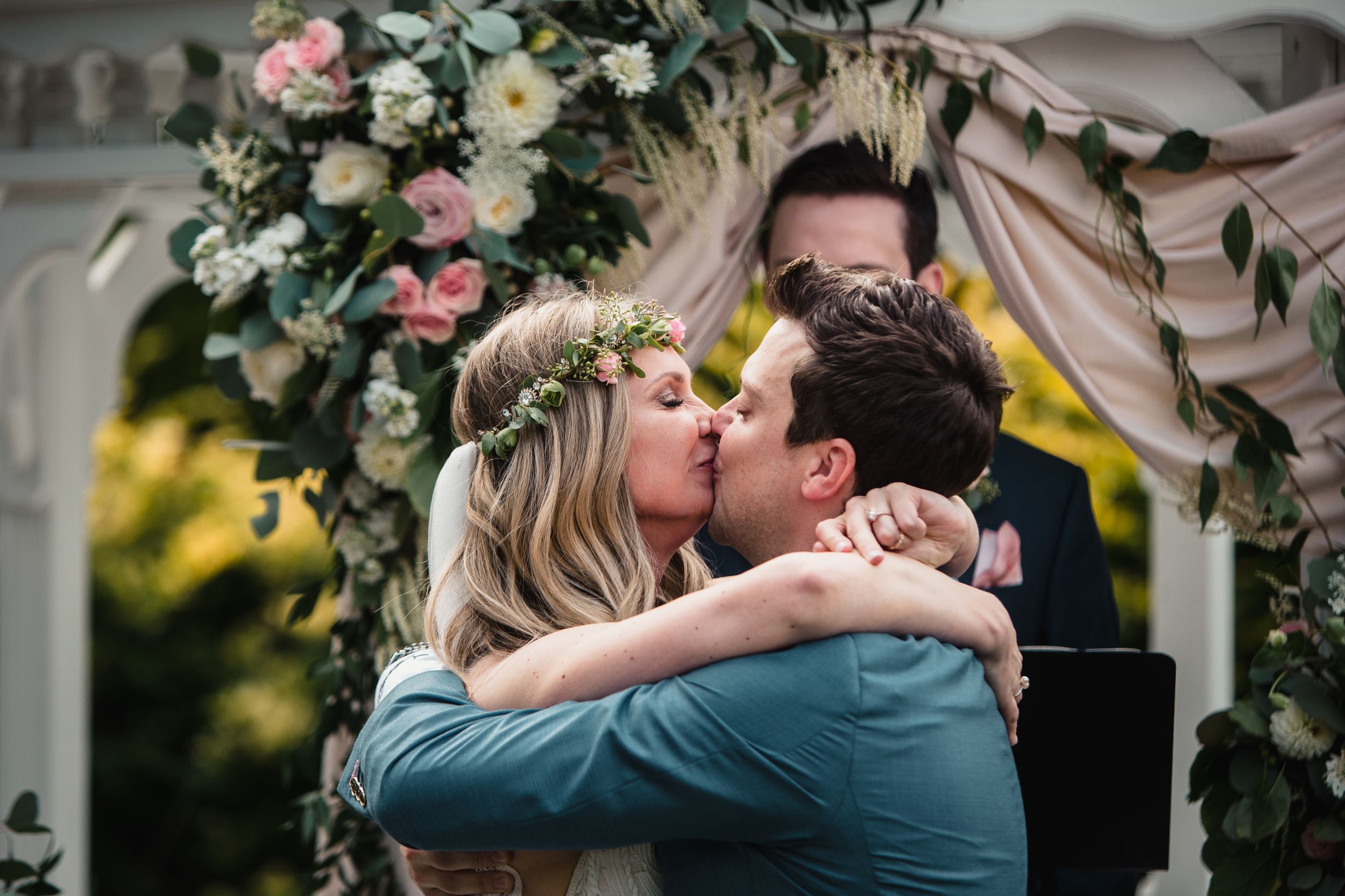 Bride and groom share first kiss at outdoor wedding ceremony under floral arch with pink roses and greenery.