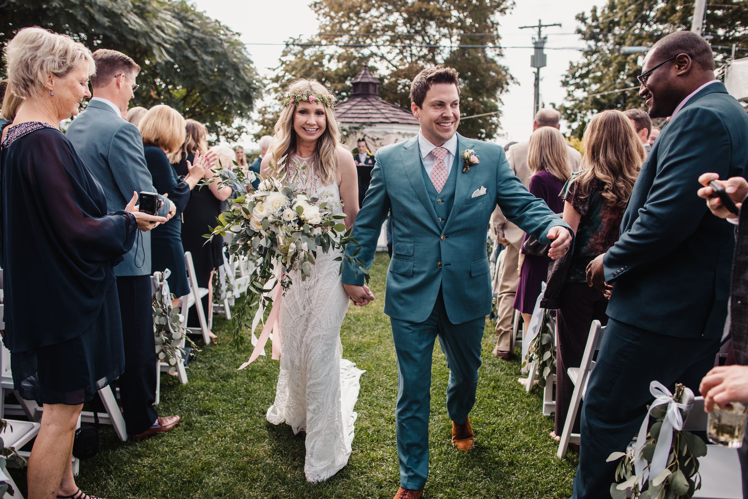Bride and groom walk down outdoor wedding aisle holding bouquet, guests applauding on both sides.