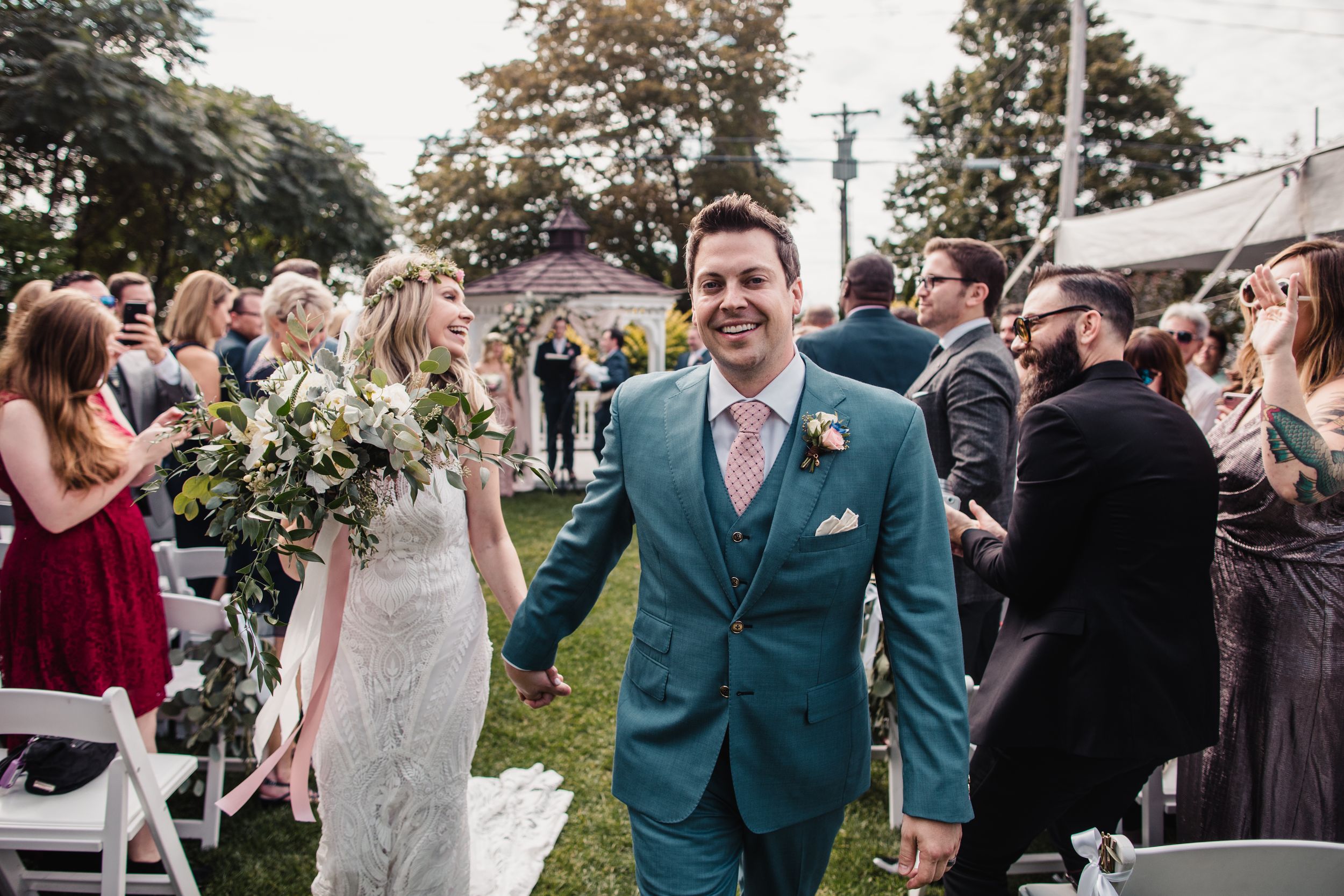 Happy newlyweds walk down the aisle after outdoor wedding ceremony, groom in teal suit, bride holding bouquet.