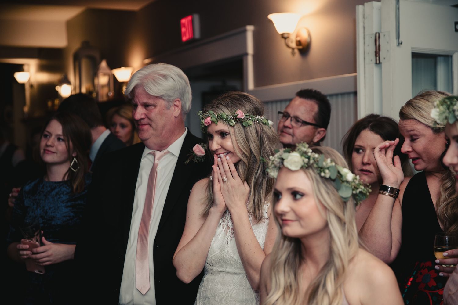 Bride wearing floral crown looks emotional surrounded by wedding guests in elegant indoor venue.