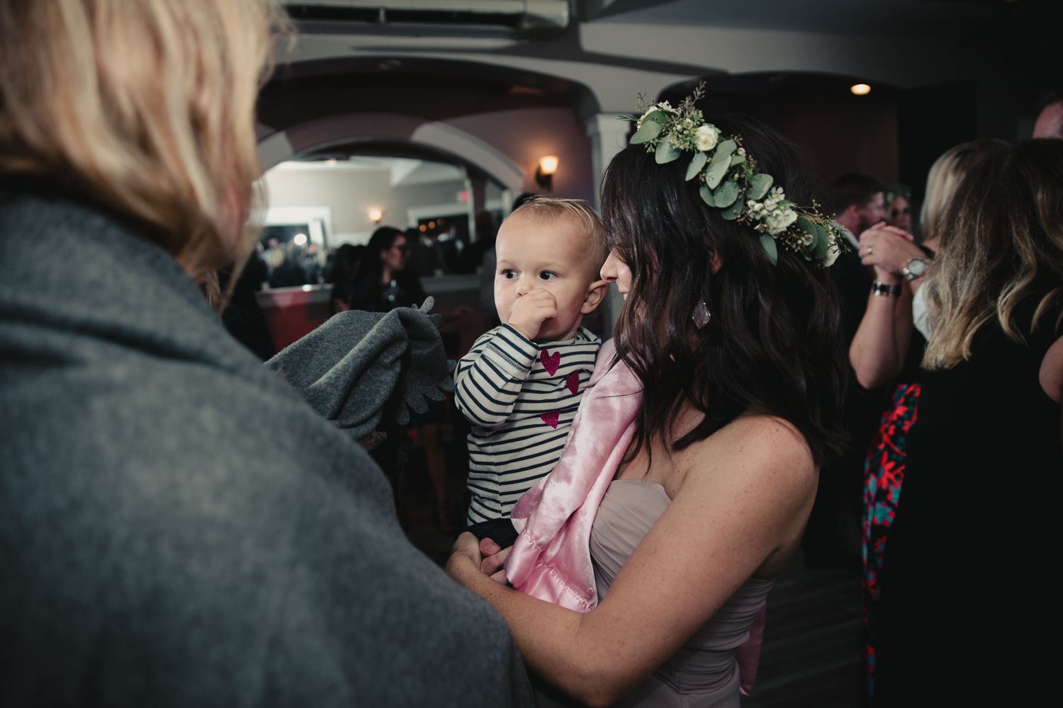 Woman wearing floral crown holds baby in striped outfit at indoor gathering.