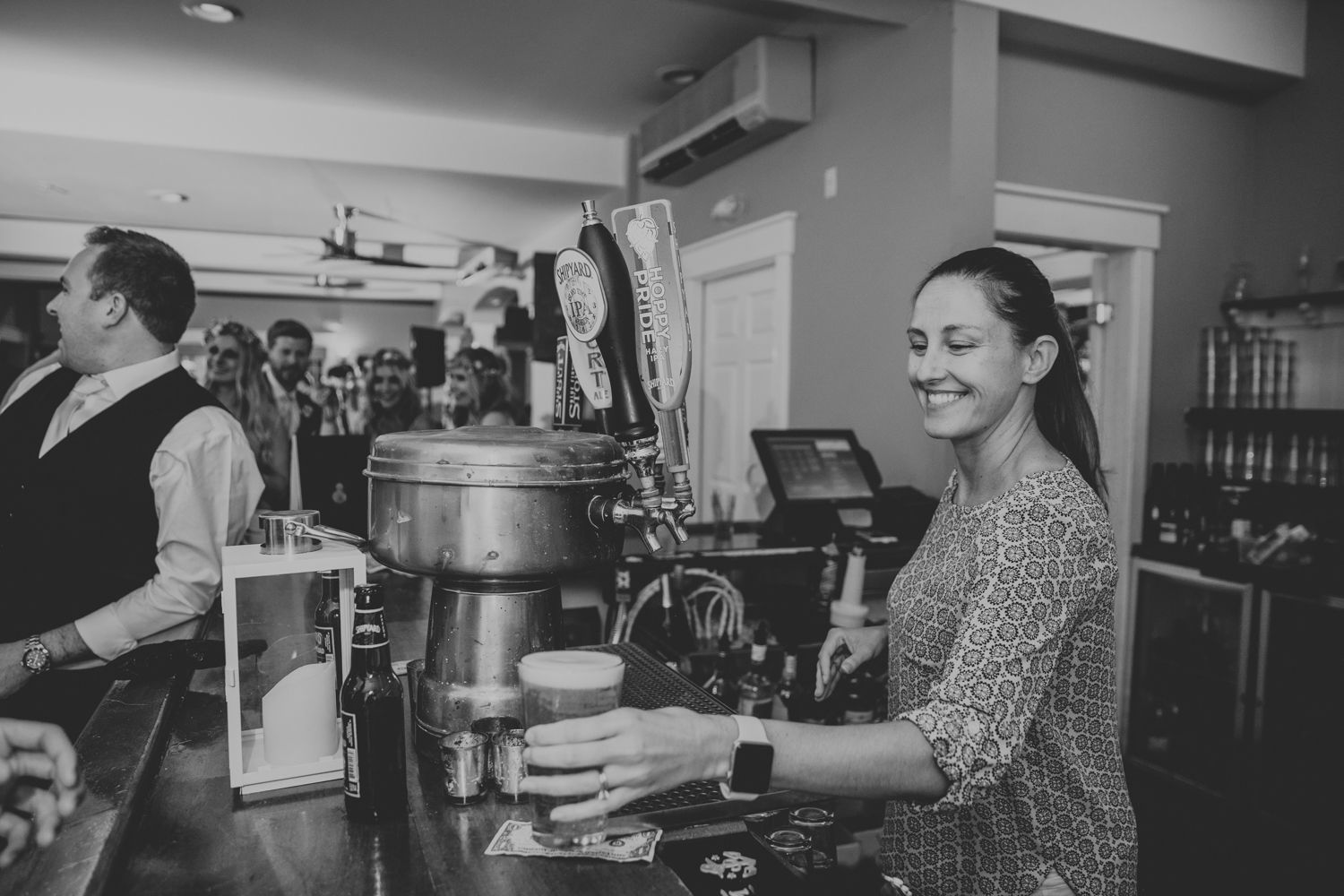 Smiling female bartender serves drinks at a busy wedding reception bar in black and white photo.