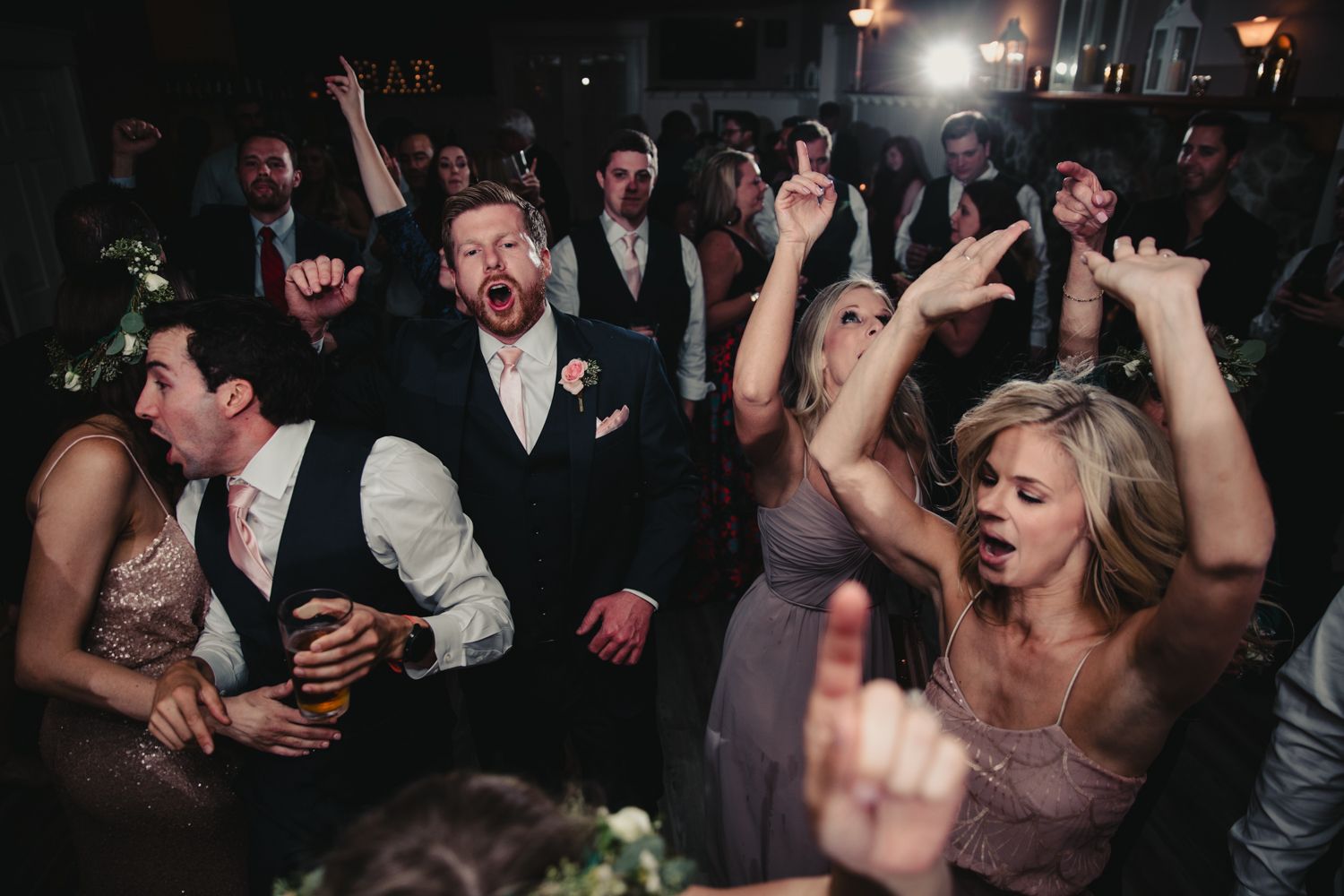 Excited wedding guests dance and celebrate with hands raised at lively indoor reception party.