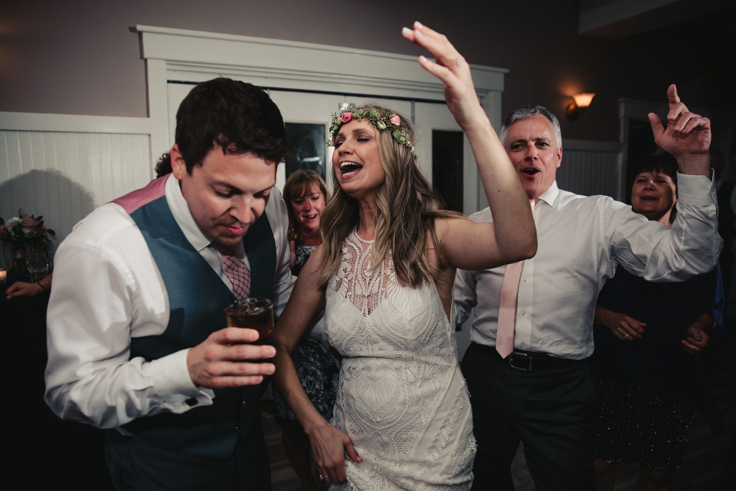 Bride in floral crown and groom dance joyfully with guests at lively wedding reception.