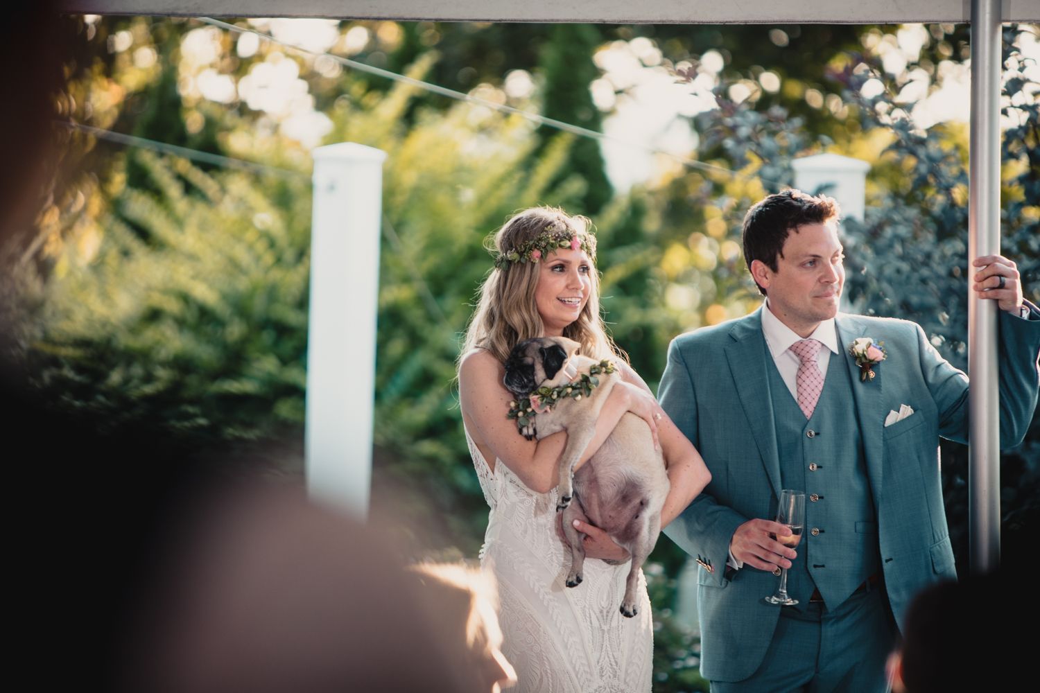 Bride holding a dog stands beside groom in blue suit at outdoor wedding ceremony under a tent.
