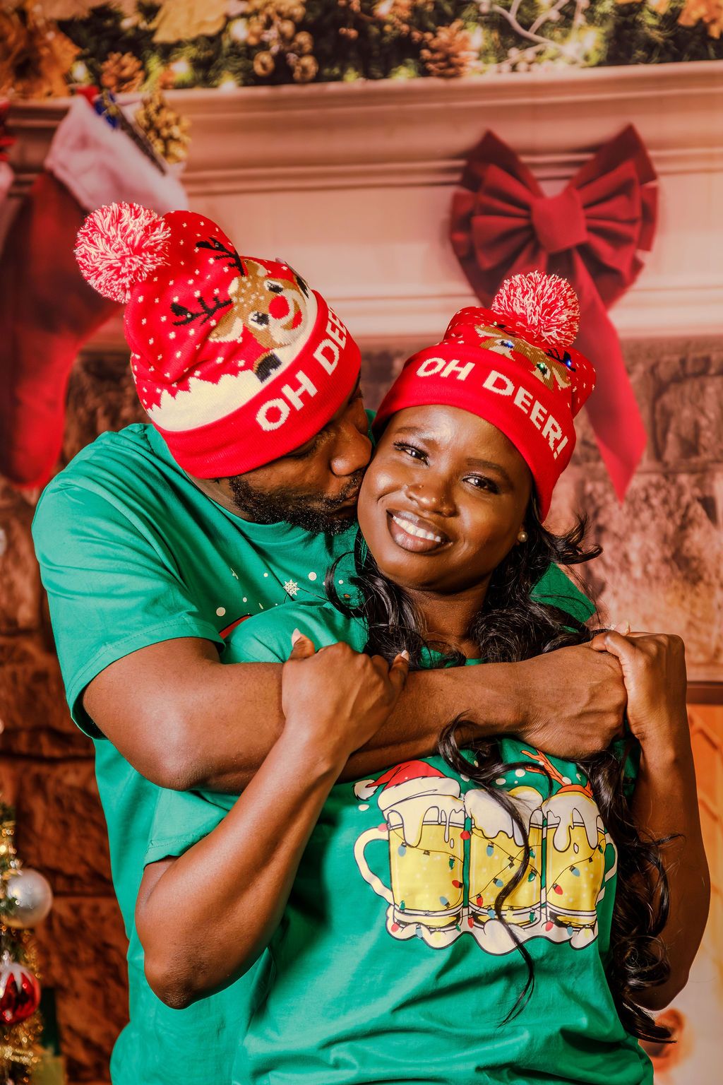 A loving Black couple in green Christmas shirts and "Oh Deer" holiday hats share a sweet embrace and kiss.