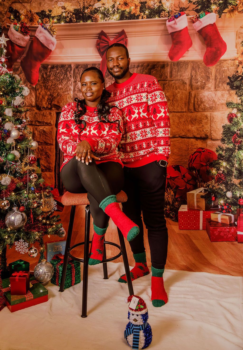 A Black couple poses in matching red reindeer ugly Christmas sweaters with festive socks near a decorated fireplace.