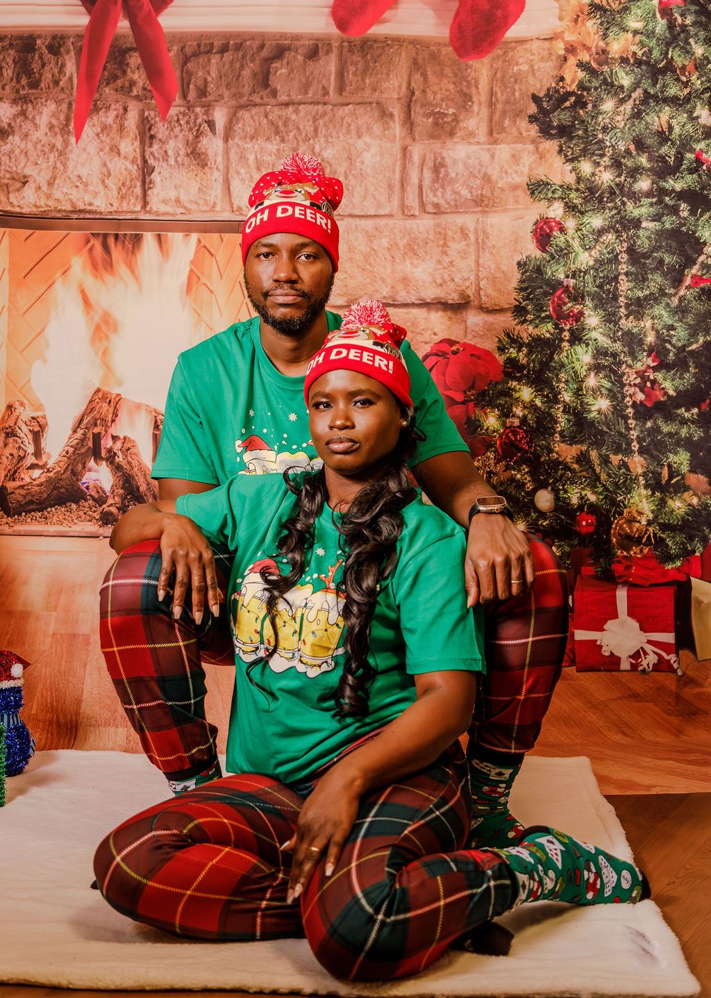 Couple in matching green Christmas pajamas and red Oh Deer beanies posing by decorated Christmas tree.