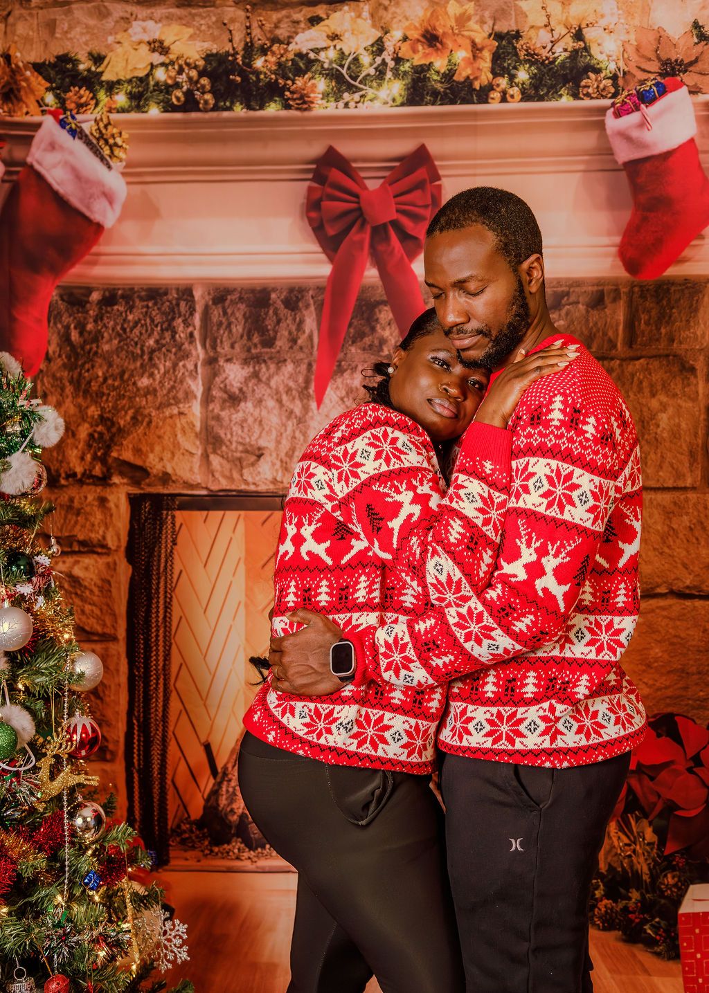 A Black couple in matching red reindeer Christmas sweaters share a tender cuddle near a festive fireplace.