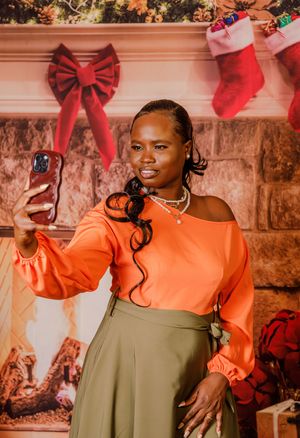 A stylish Black woman in an orange top and olive skirt takes a selfie near a Christmas fireplace backdrop.