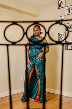 A Black woman in a stunning teal sequin off-shoulder gown with a slit poses behind a decorative iron railing.