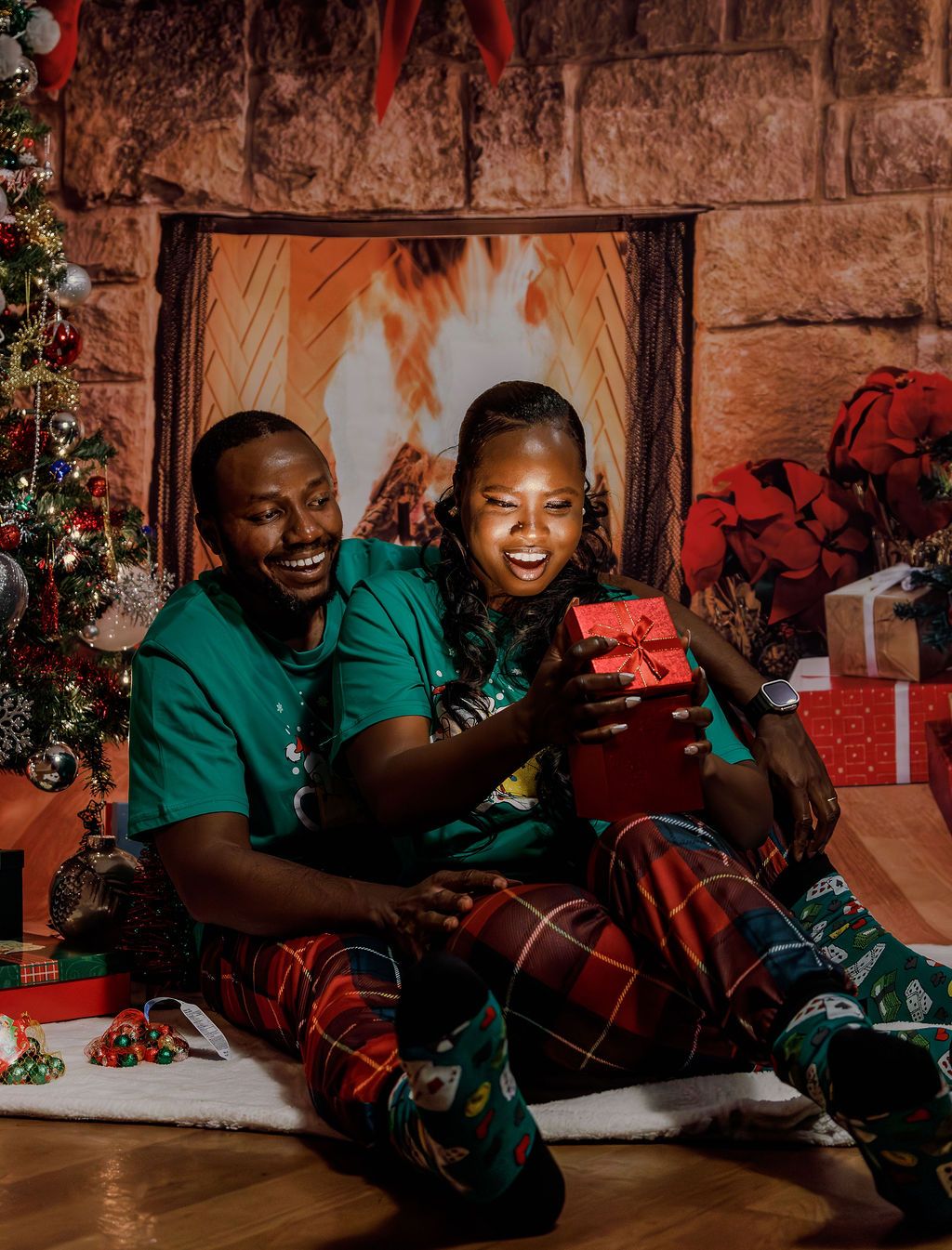 A joyful Black couple in matching Christmas pajamas sit together opening a red gift box near a decorated tree.