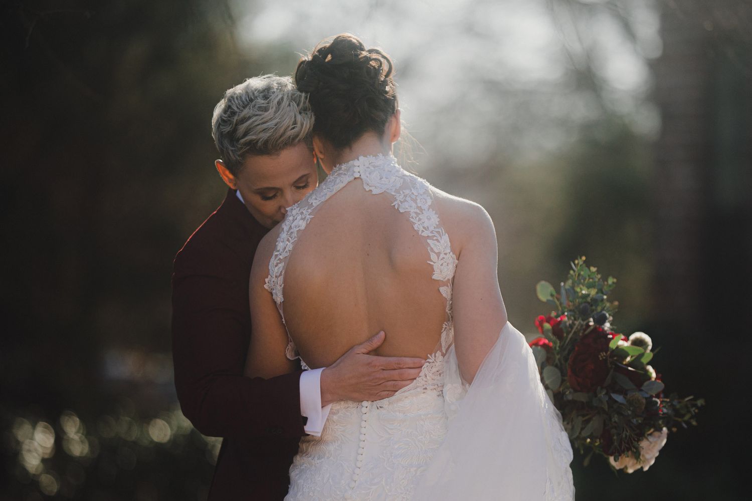 Two brides embrace outdoors, one in a backless lace wedding gown holding a burgundy bouquet, bathed in warm sunlight.