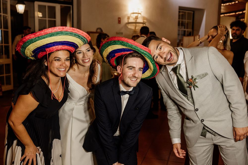 Four people wearing colorful sombrero hats pose together at a wedding reception, two in formal attire smiling joyfully.