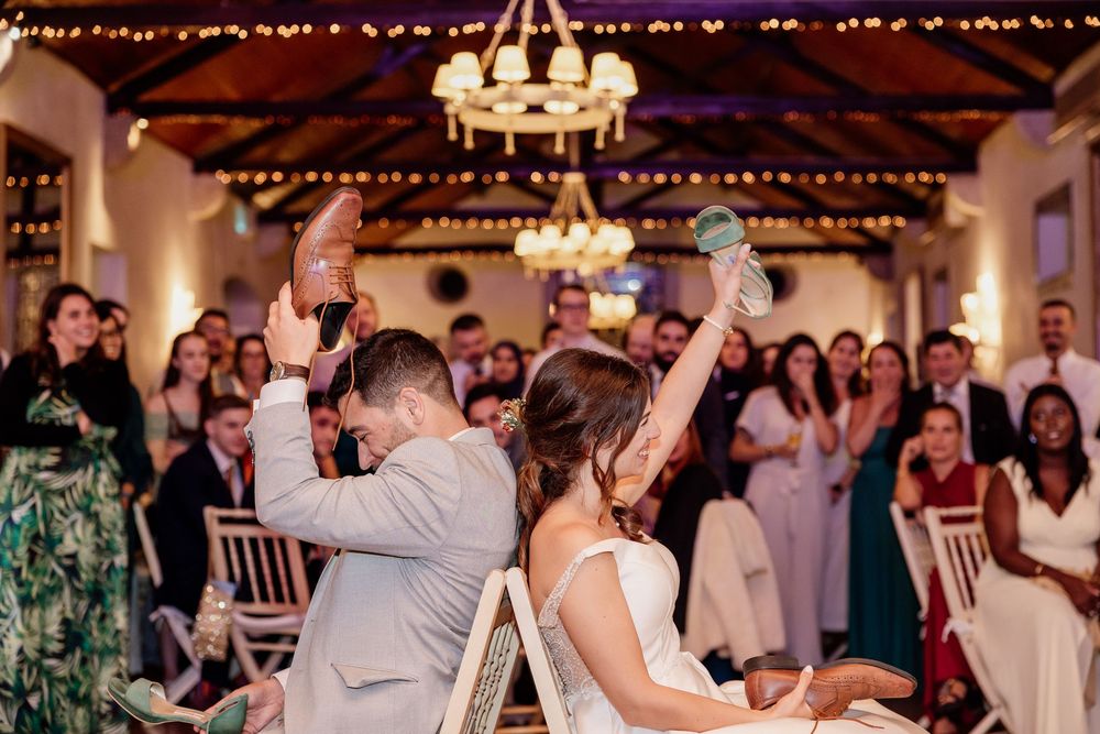 Bride and groom raise shoes during fun wedding shoe game while guests cheer in elegant reception venue.