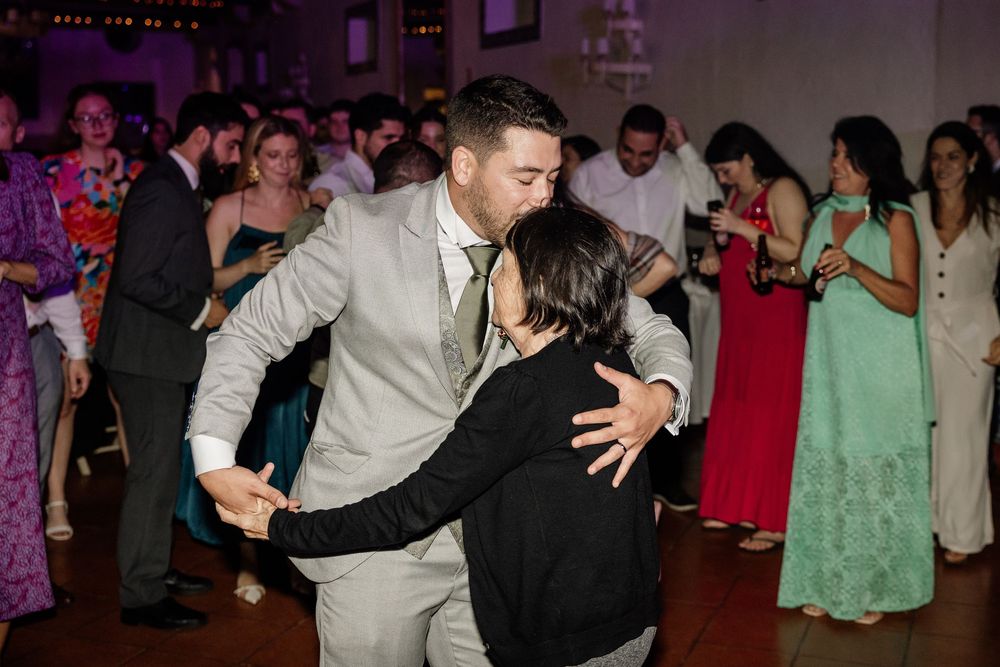 A groom in a light gray suit dances with a guest at a lively wedding reception.
