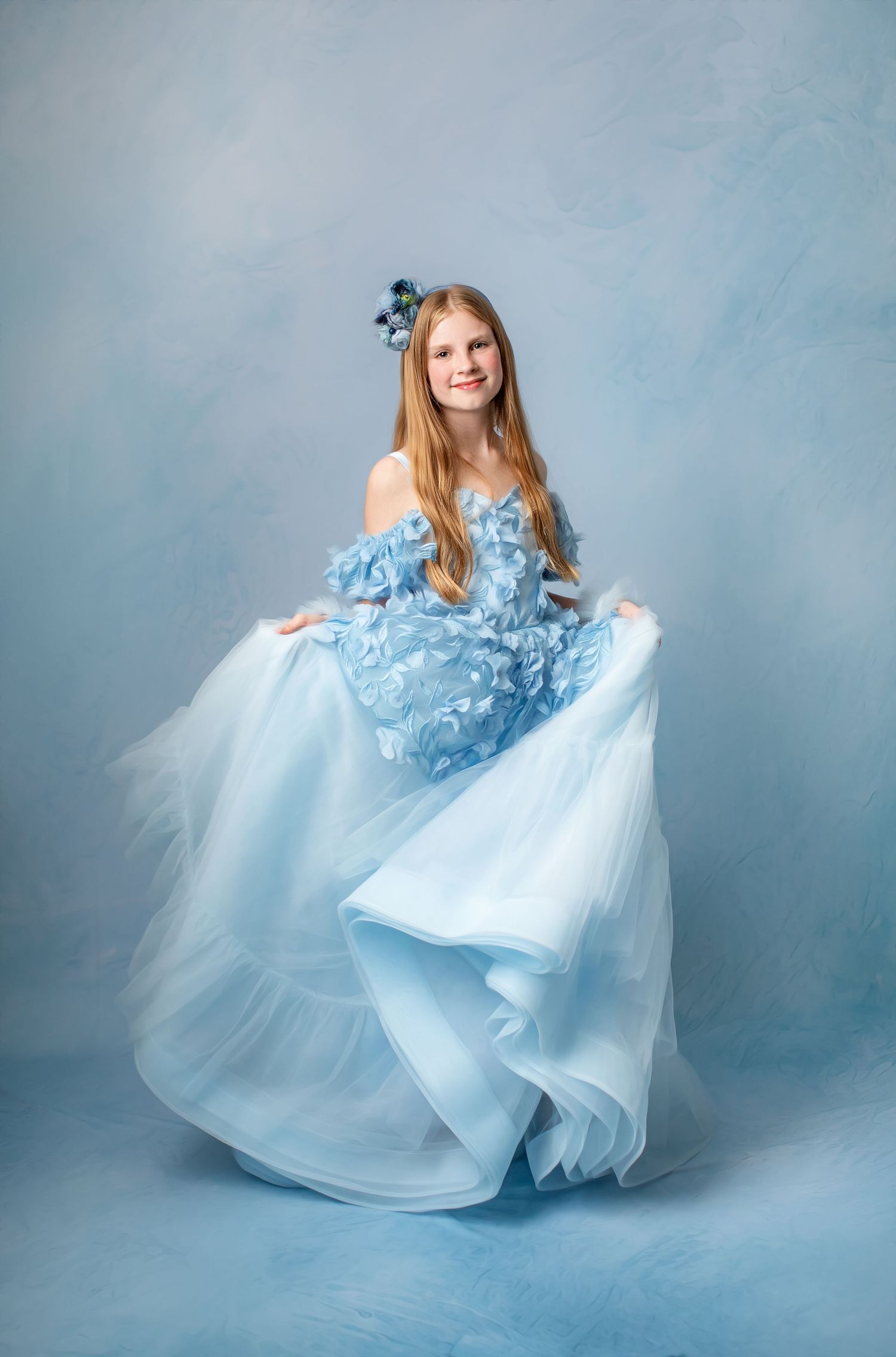 Ethereal portrait in flowing ice blue layered tulle gown with floral details against pale backdrop.