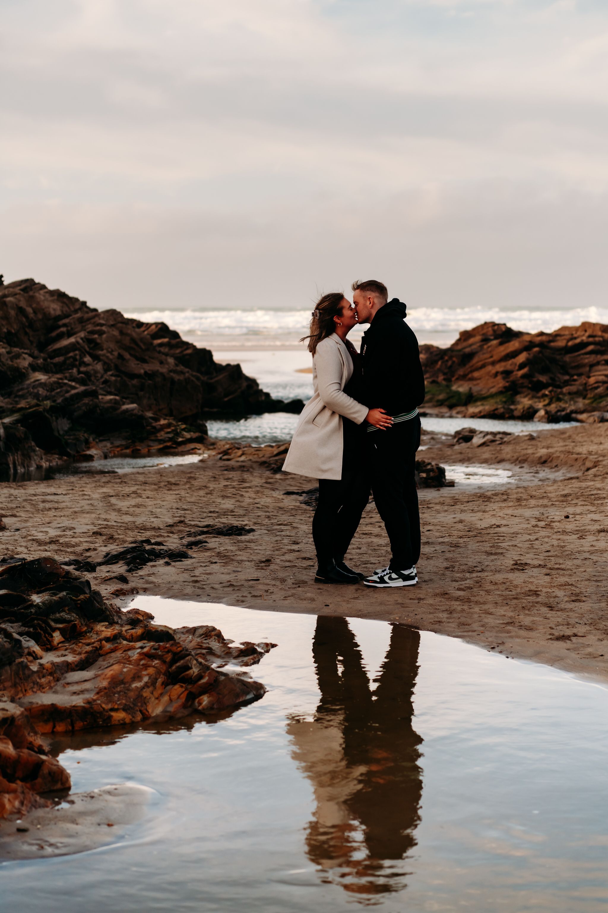 A couple embraces on a rocky beach at sunset with waves crashing against the shore and their reflection in tidal pools.