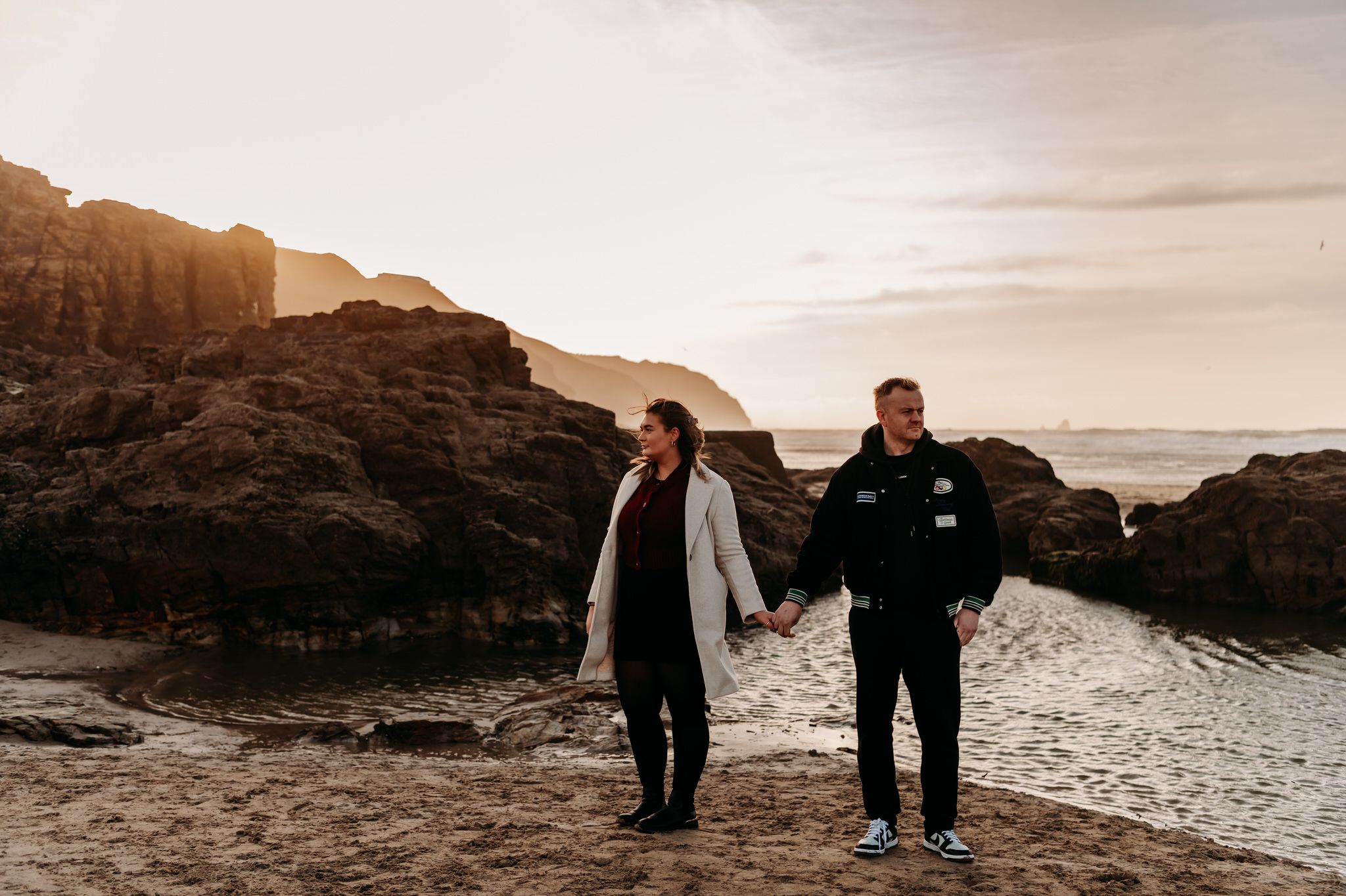 Two figures stand silhouetted against a dramatic coastal landscape at sunset with rocky cliffs and ocean views.