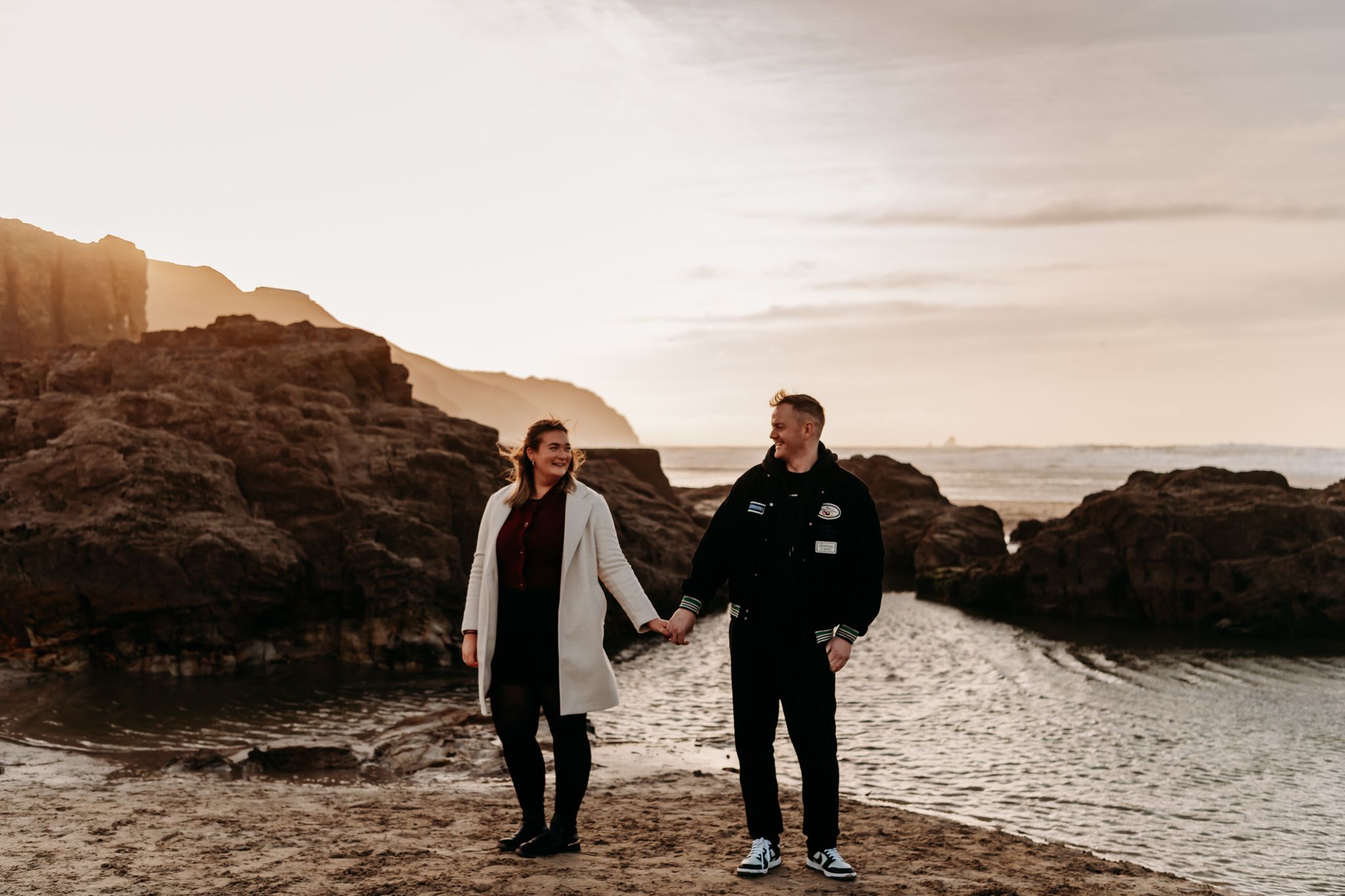 Two people walk along a rocky coastal beach during sunset with dramatic cliffs in the background.