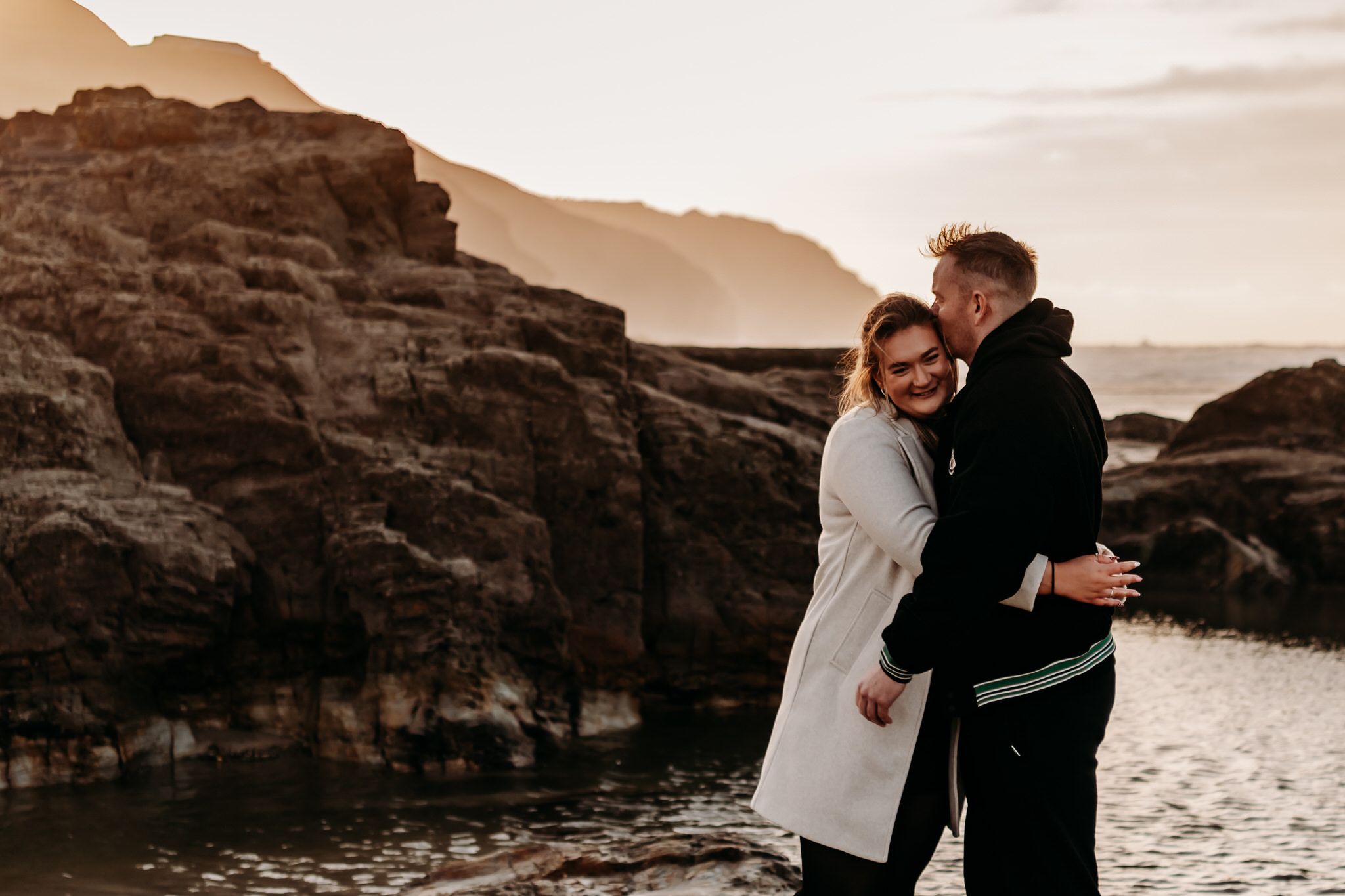 A couple embraces on a rocky coastline at sunset with mountains in the background creating a romantic silhouette.