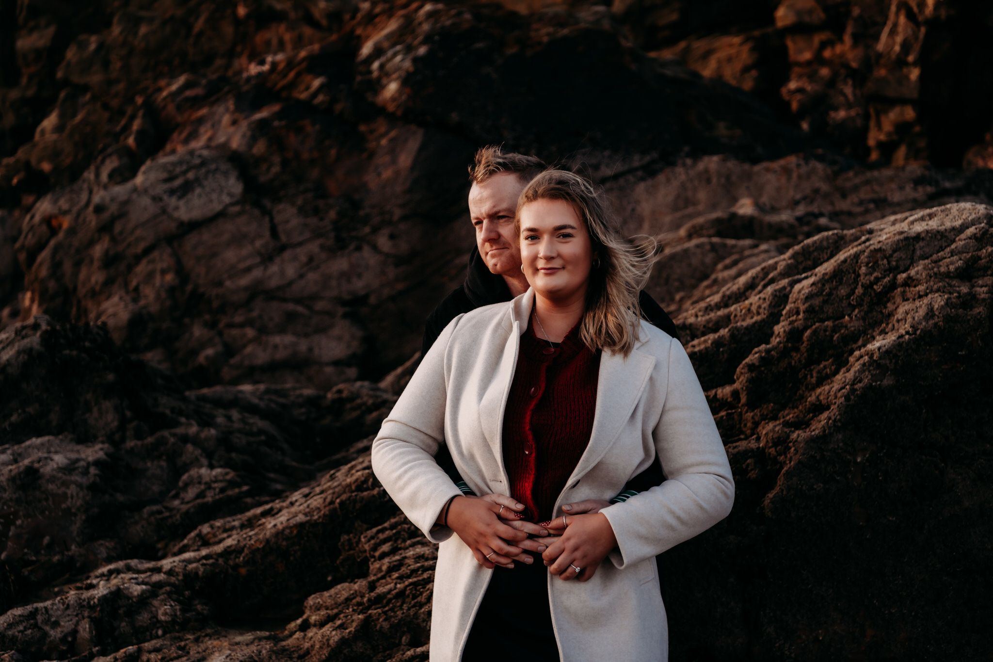 A couple embraces while standing against a rocky cliff backdrop during an outdoor photoshoot.