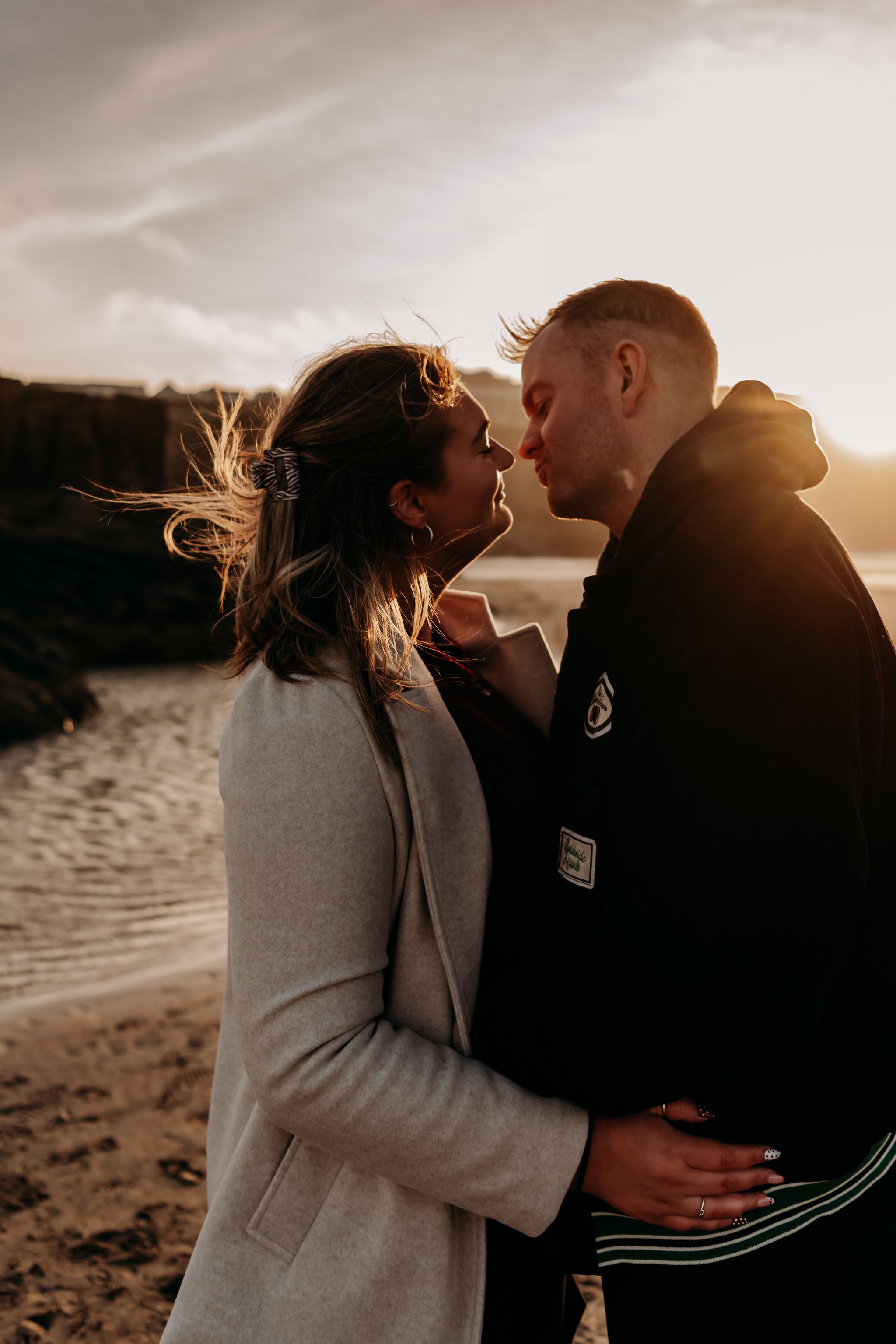 A couple shares an intimate moment at sunset on a beach while wearing casual winter clothing.
