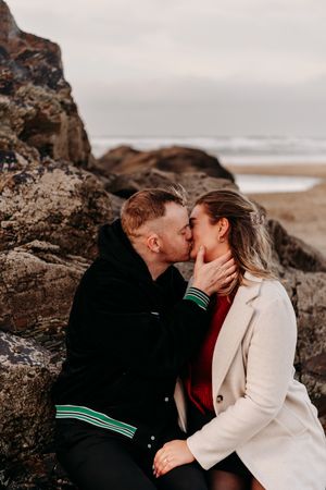 A romantic coastal photoshoot featuring a couple sitting on rocky cliffs by the ocean at sunset.