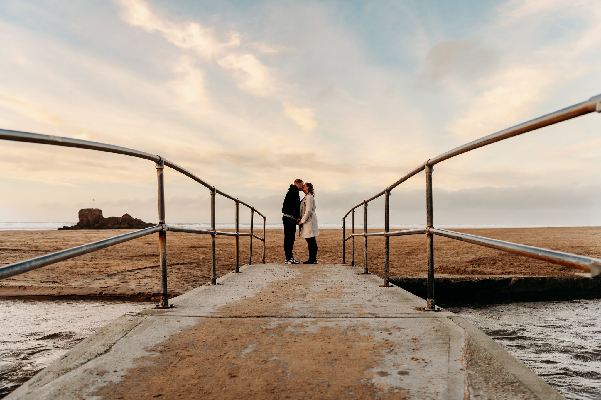 Two people stand together on a wooden pier overlooking a serene lake during golden hour sunset.