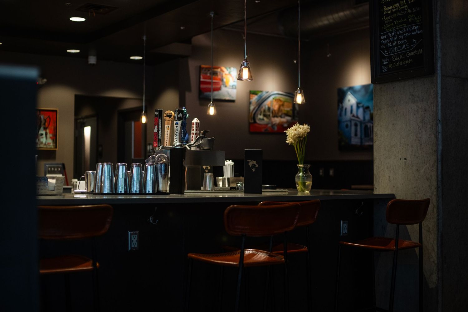 Dimly lit bar interior with orange stools, pendant lights, beer taps, and colorful artwork on dark walls.