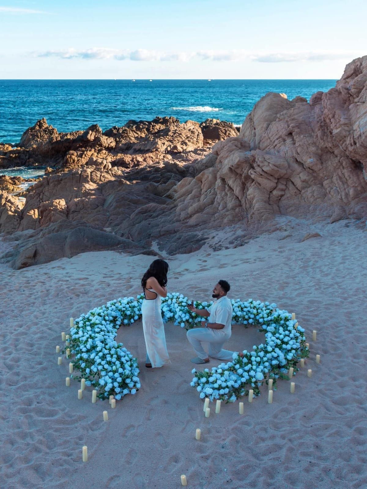 Romantic beach proposal with heart-shaped floral arrangement of white flowers and candles by rocky shoreline at dusk.