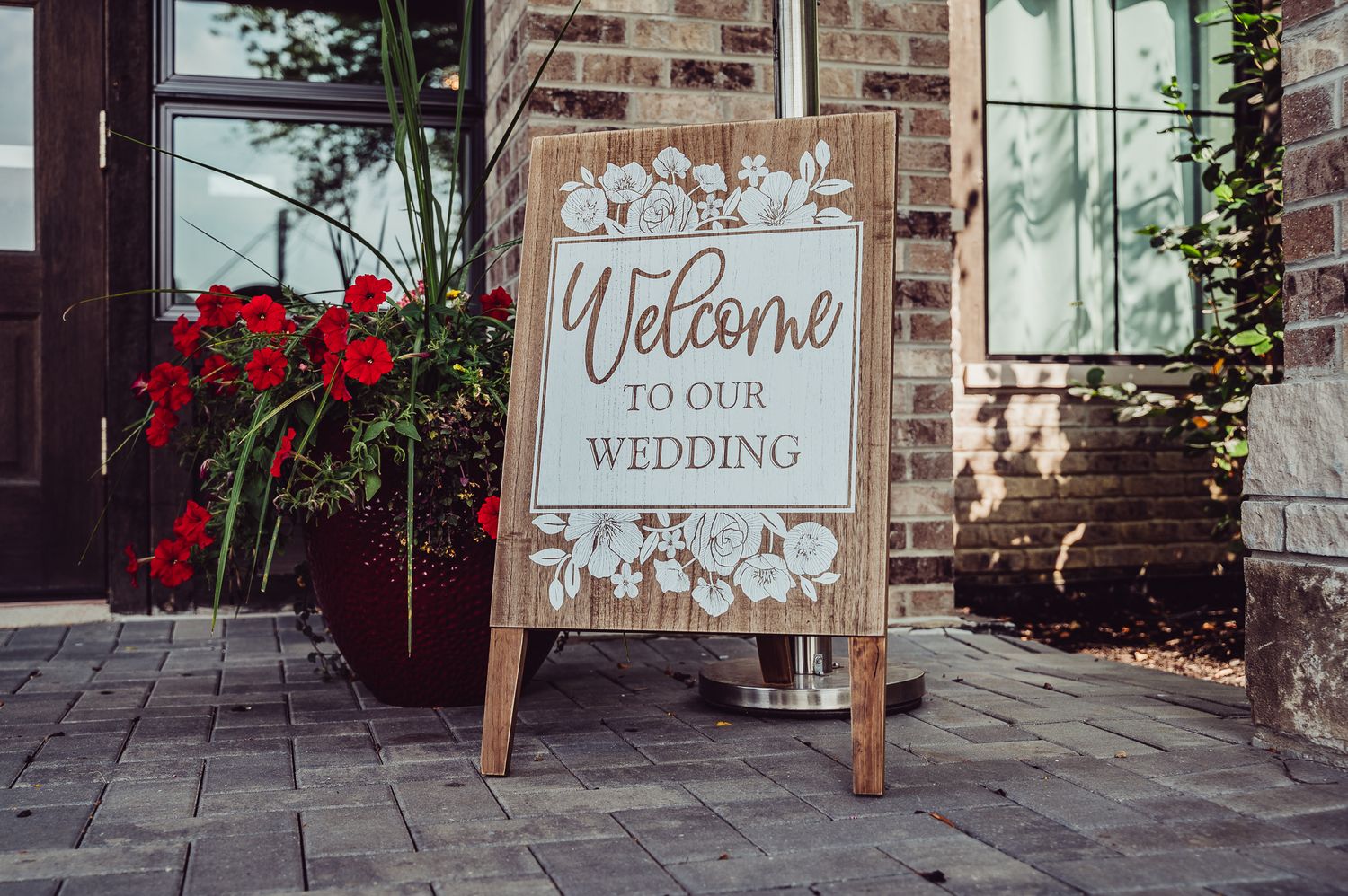 Wooden A-frame welcome to our wedding sign on brick patio with red flowers and greenery.