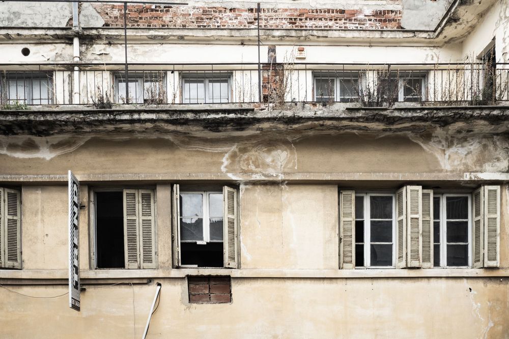 Old weathered building facade with peeling paint and open shuttered windows on a decaying European style structure.