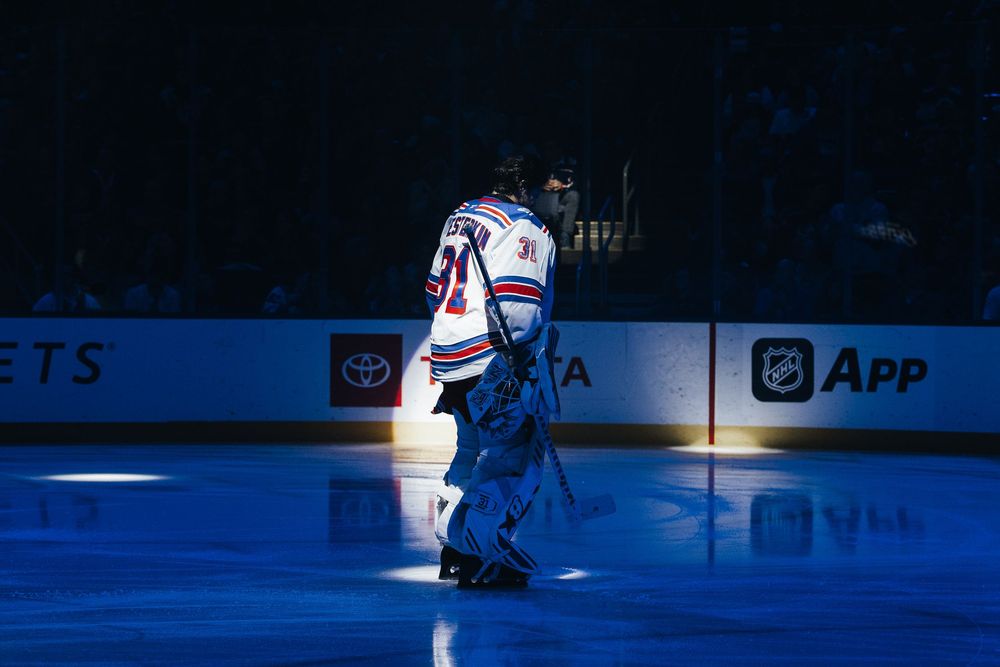 A hockey goalie stands in the spotlight on a blue-lit ice rink during a pre-game introduction.