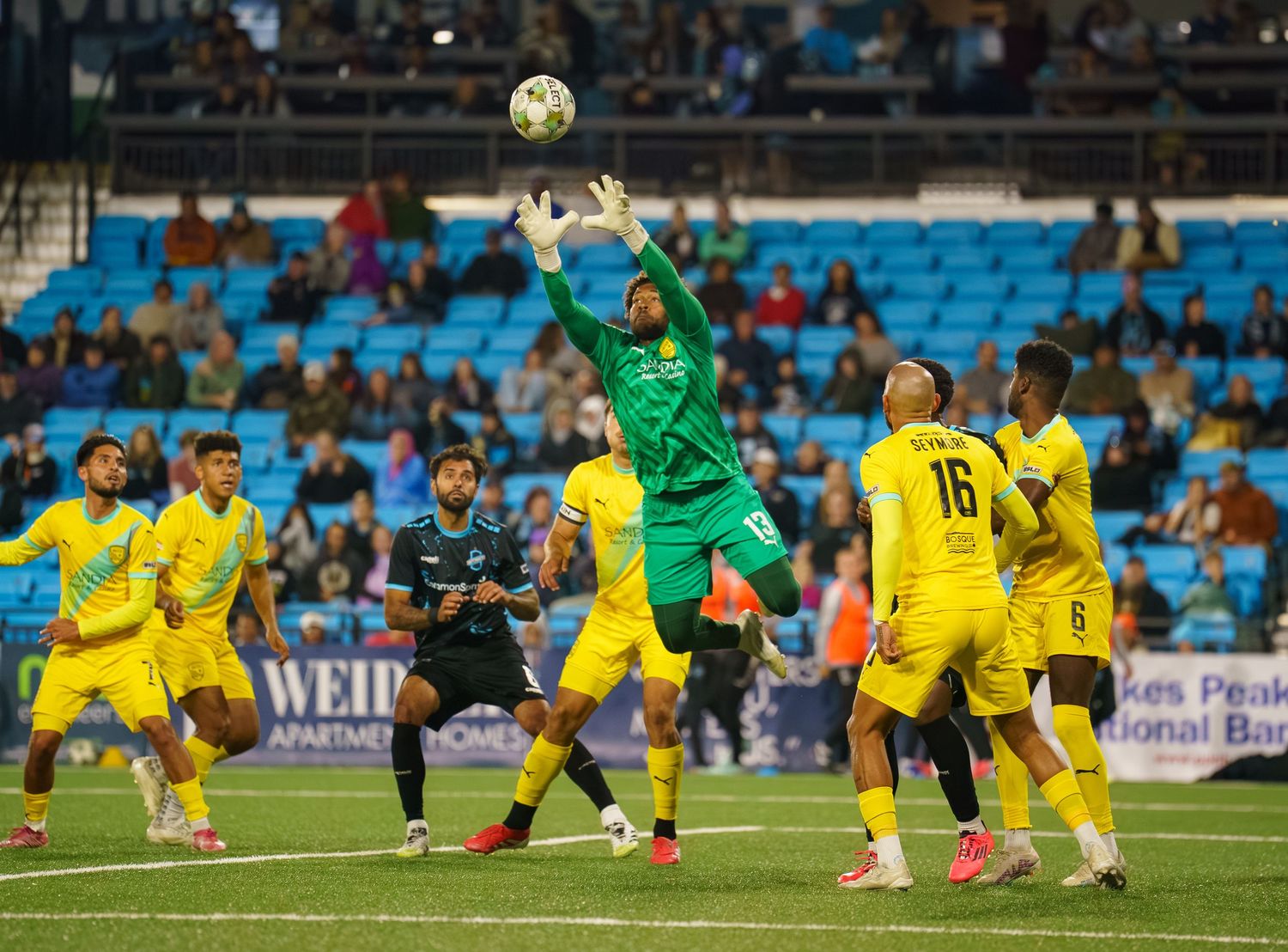 Goalkeeper in green jersey leaps to make a save during soccer match against players in yellow uniforms.