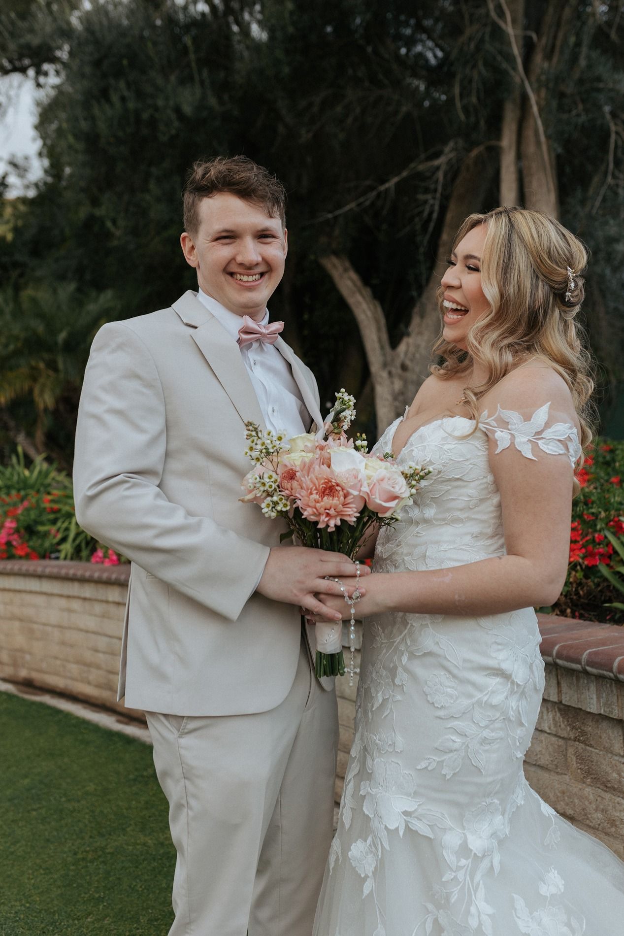 A newlywed couple shares a joyful moment in a garden setting with pink floral bouquet and light-colored attire at Singing Hills Golf Resort.